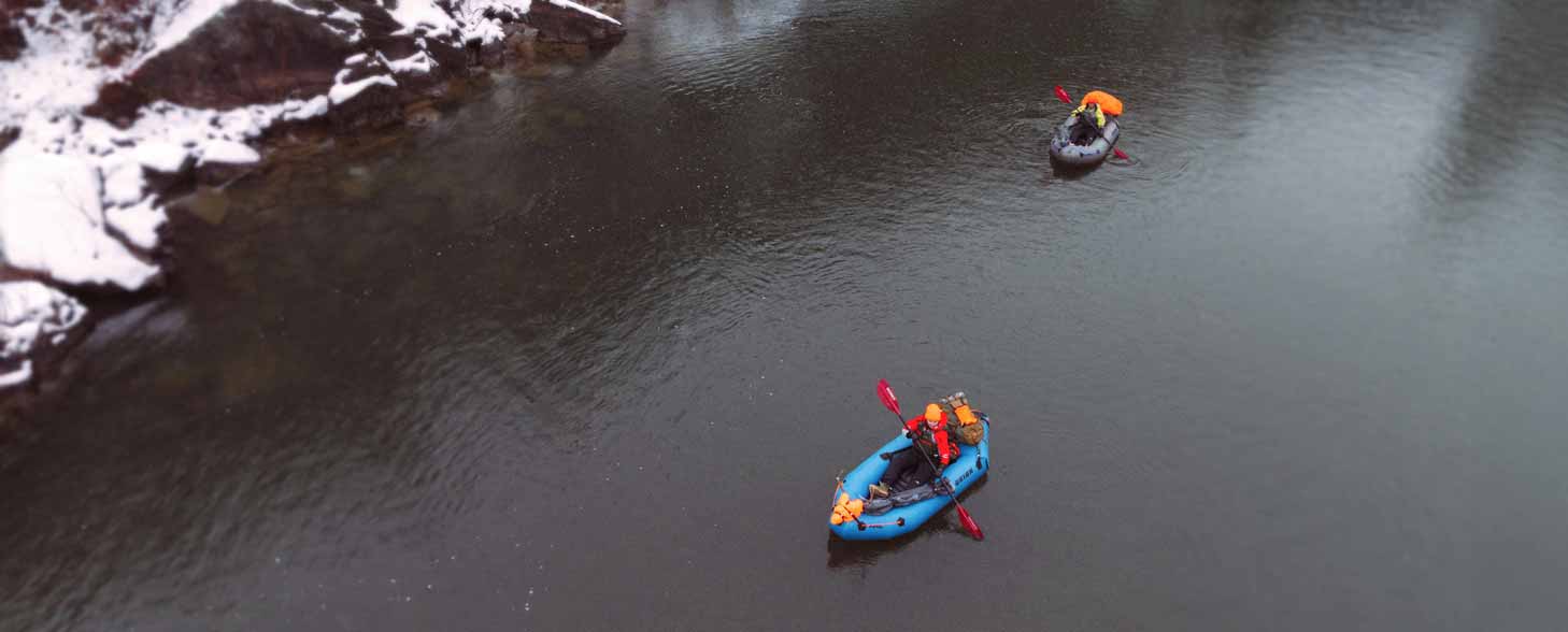 An aerial via of two hunters paddling their packrafts down a river in winter.