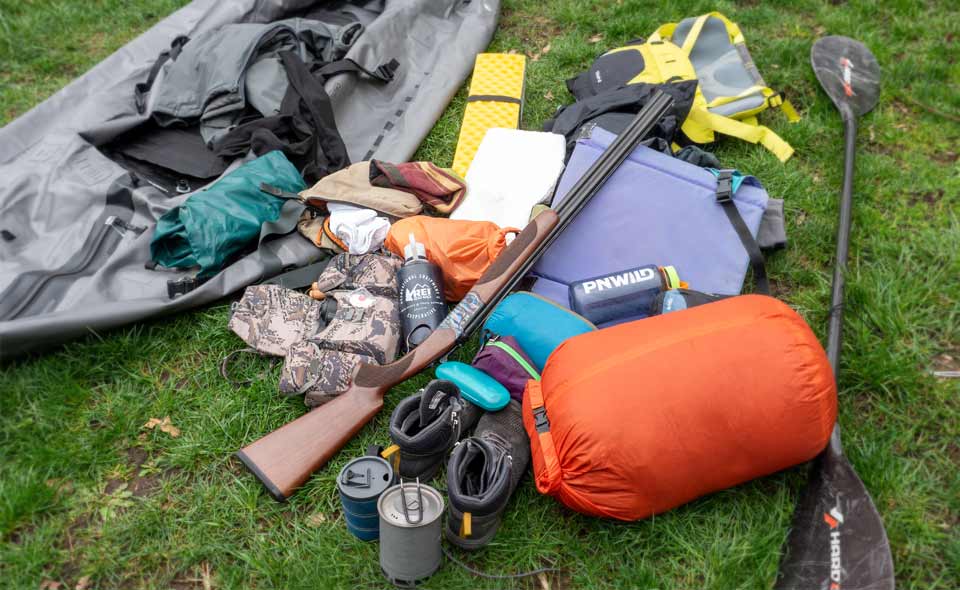 The essential accessories for hunting with a packraft are laid out on the ground next to a deflated Orion packraft. 
