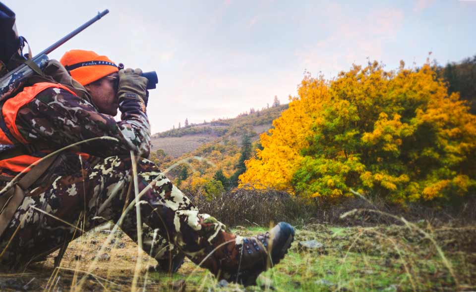 A hunter in camo attire and backpack sits and looks through binoculars.