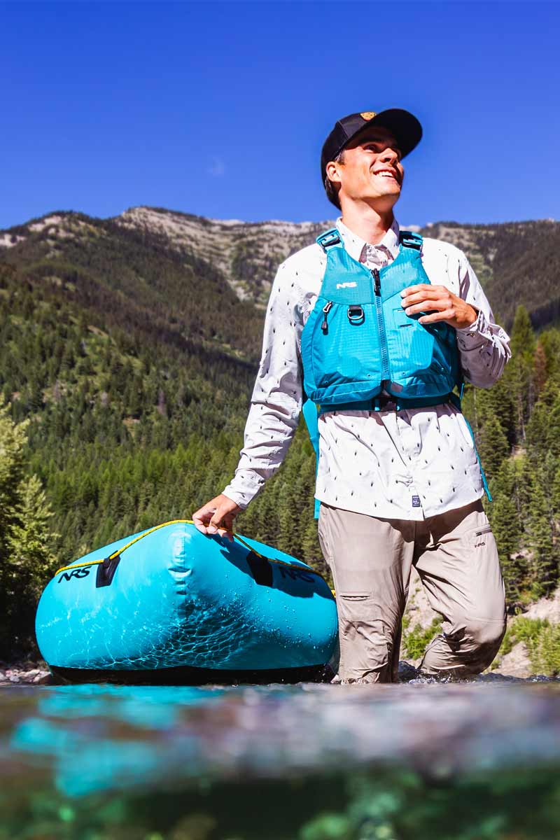A man walks in knee-deep water while pulling his NRS packraft with him.