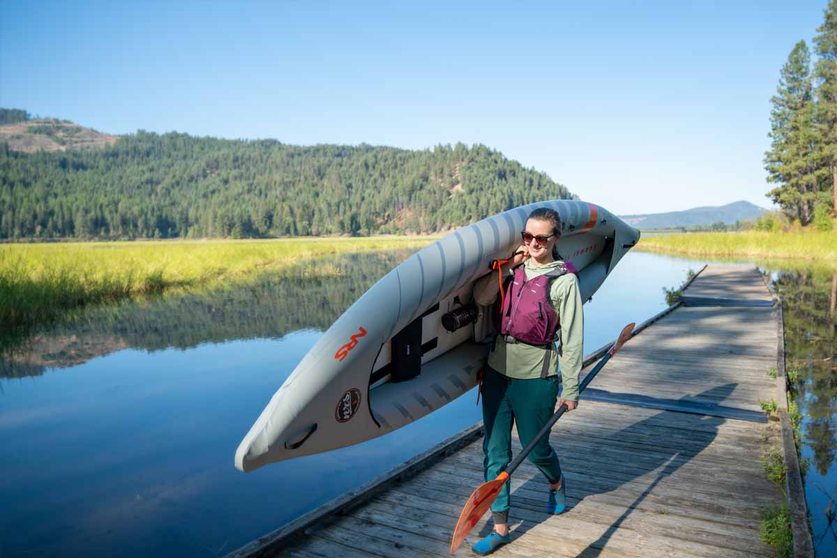 A woman happily carries the Akamai 110 inflatable kayak over her shoulder down a riverside dock.