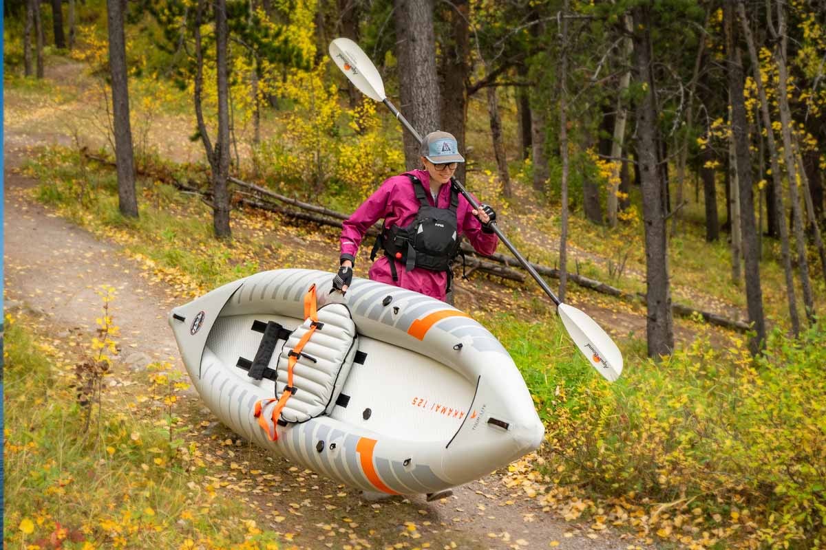 A woman carries the Akamai 125 inflatable kayak down a fall trail with her paddle slung over her shoulder.