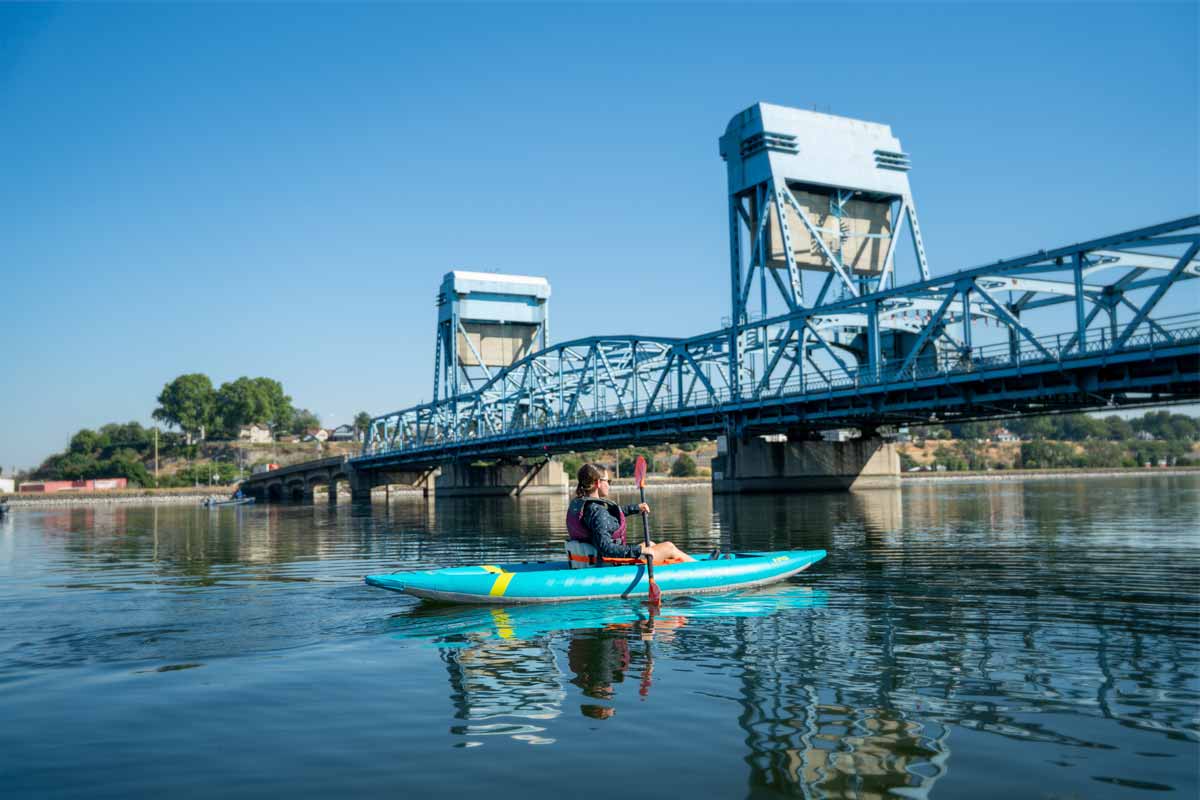 A woman paddles the Kaholo 110 kayak towards a large bridge overpass.