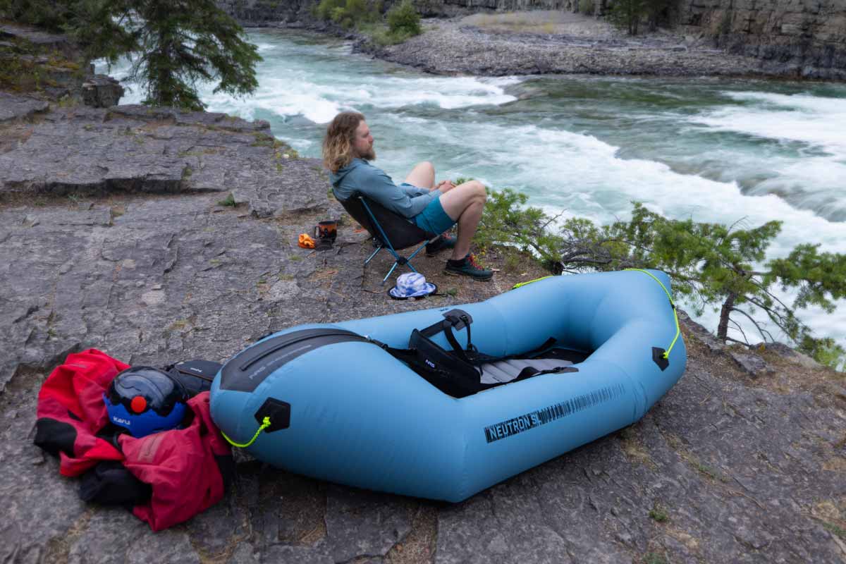 A man relaxes in a camp chair next to the Neutron SL Packraft while overlooking a river.