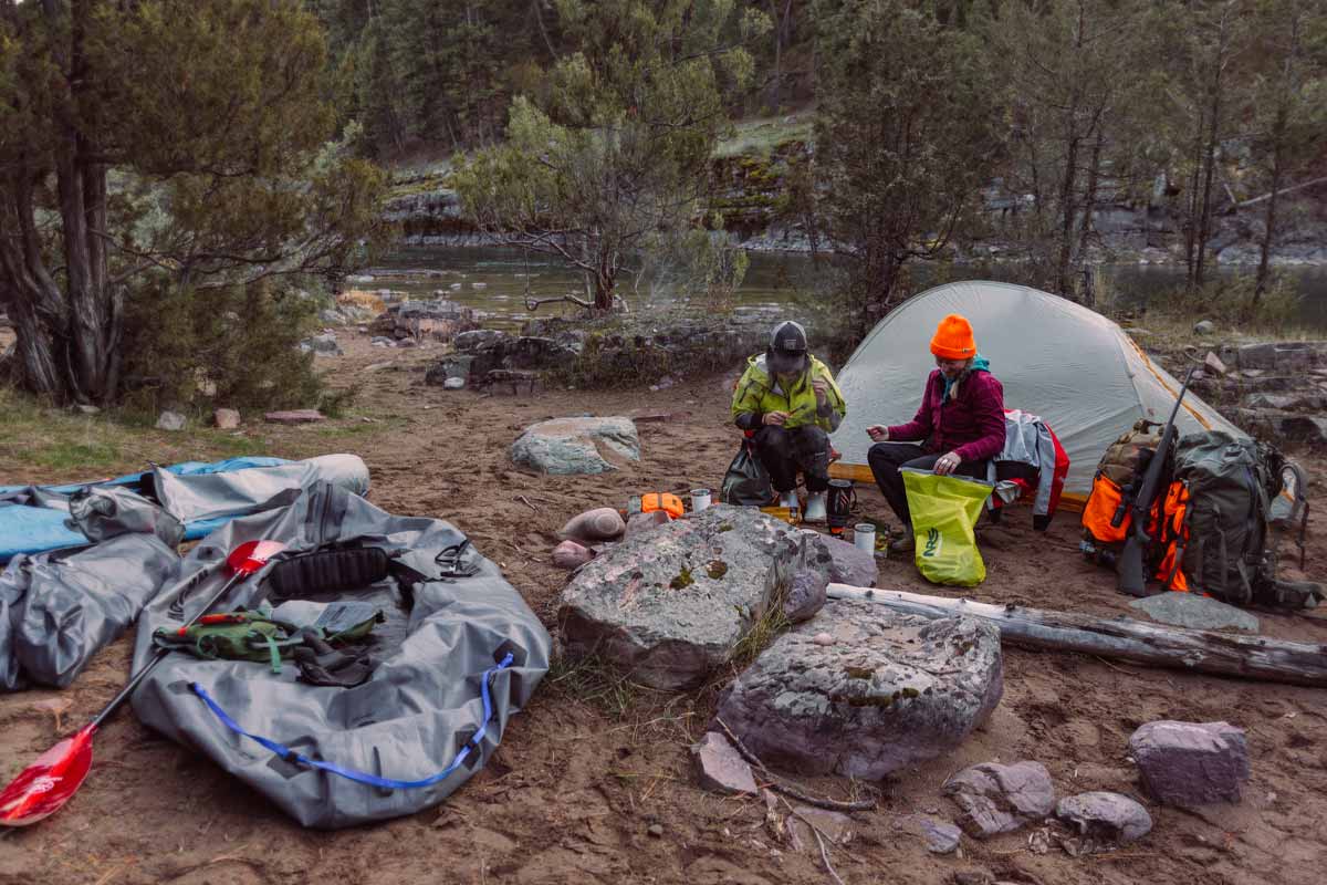 Two women relax in camp chairs at their riverside campsite surrounded by hunting gear.