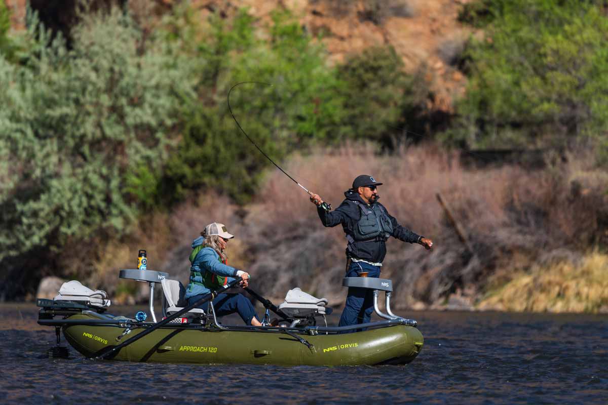 A man casts his fishing line from the bow of the Orvis Approach 120 while a woman rows the oars.