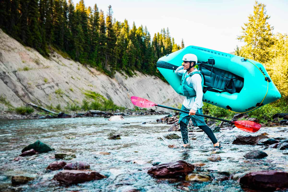 A man carries the Pulsar packraft through a shallow river crossing.