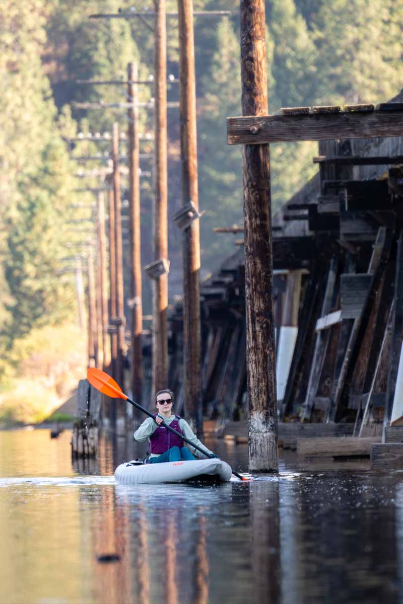 A woman paddles parallel the large wooden trusses of a train bridge in the Akamai 110 kayak.