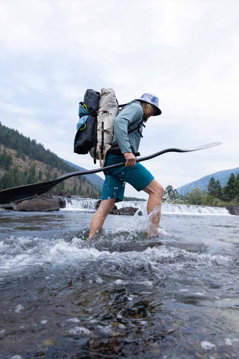 Man hikes through a shallow river toting a kayak paddle in hand and a packraft stowed on his back.