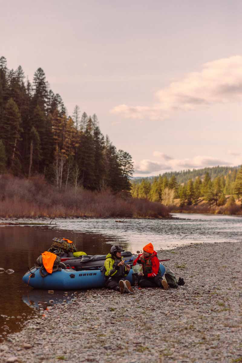 Two women lean against their Orion Packrafts while eating a snack on the riverbank.